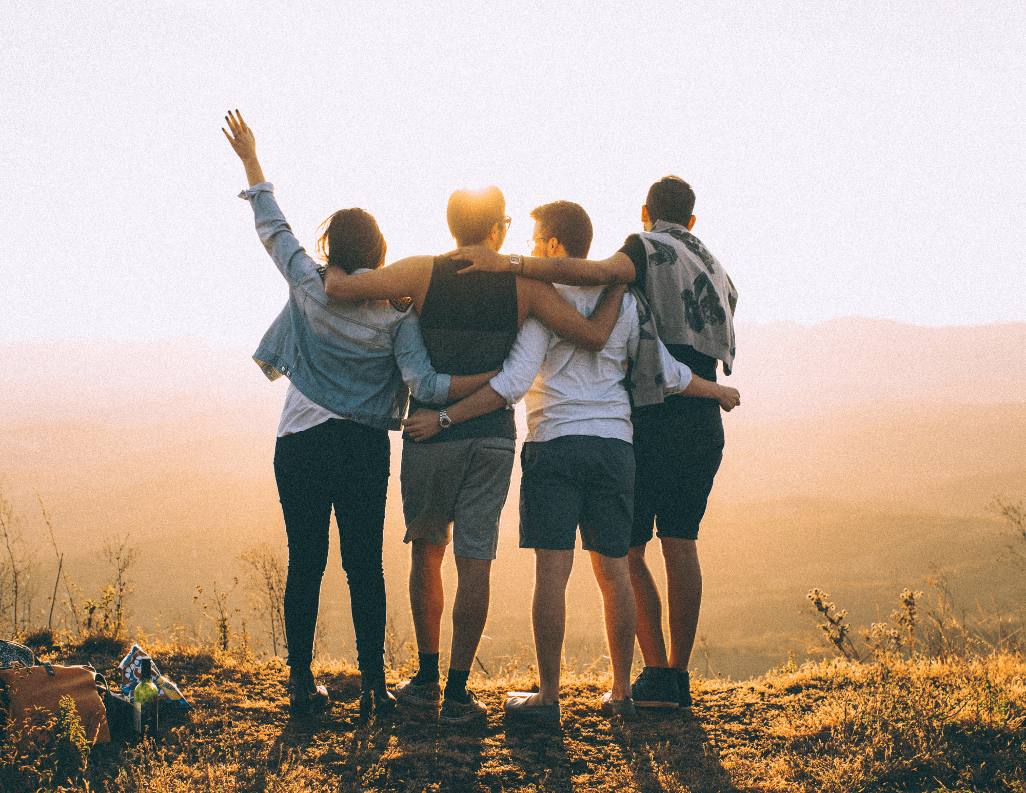 Vus de dos, quatre amis debout sur une colline, bras dessus, bras dessous ou avec un bras tendu, contemplent le soleil à l’horizon.