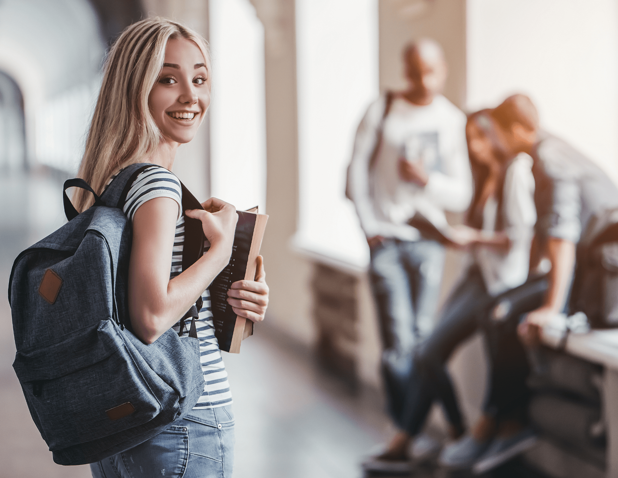 Une jeune femme blonde souriante tient son sac à dos et des manuels scolaires, debout dans un couloir d’école. En arrière-plan, trois élèves sont assis.