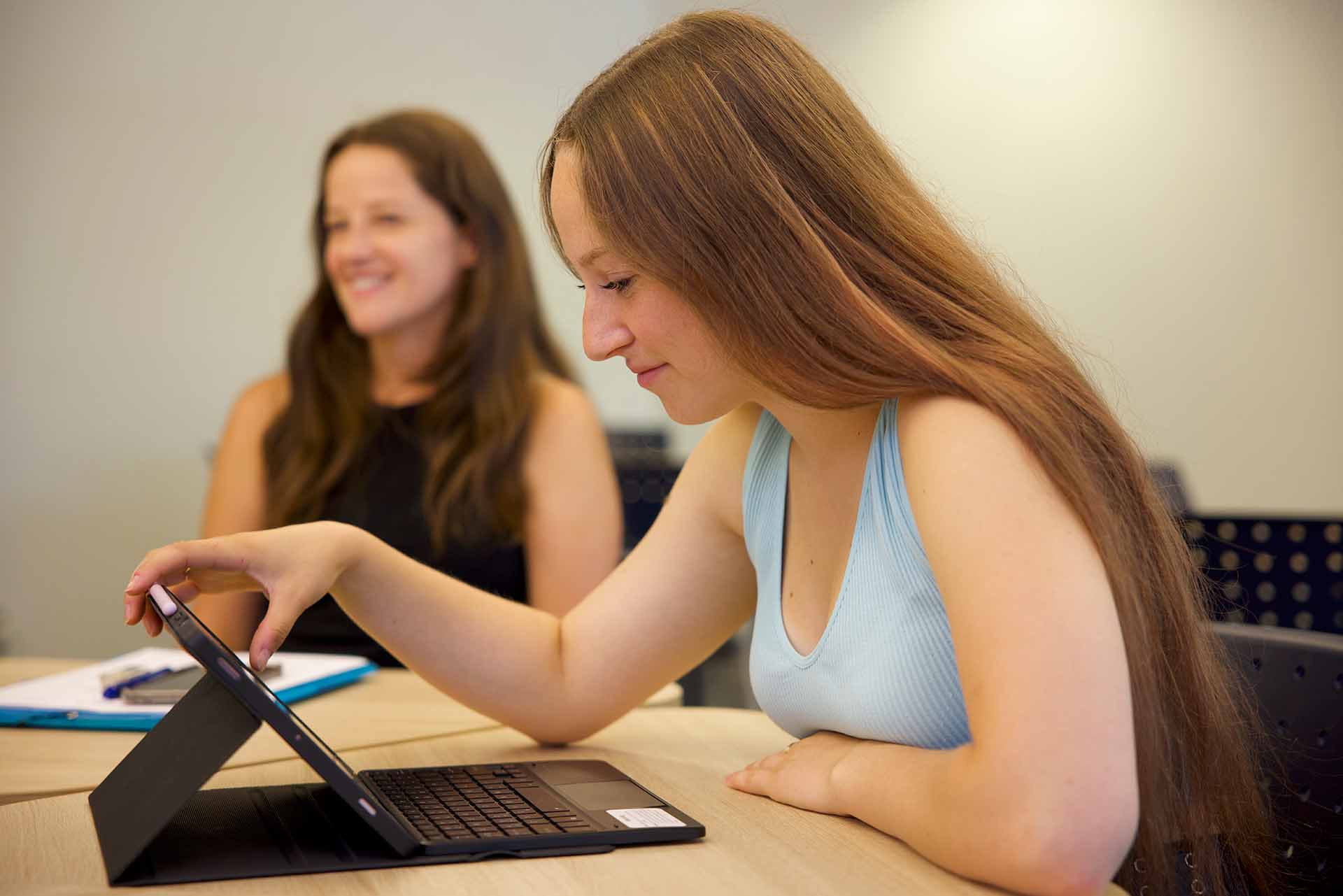 Deux jeunes femmes sont assises dans une salle de classe. L’une sourit à l’arrière-plan, tandis que l’autre regarde un ordinateur portable en souriant.