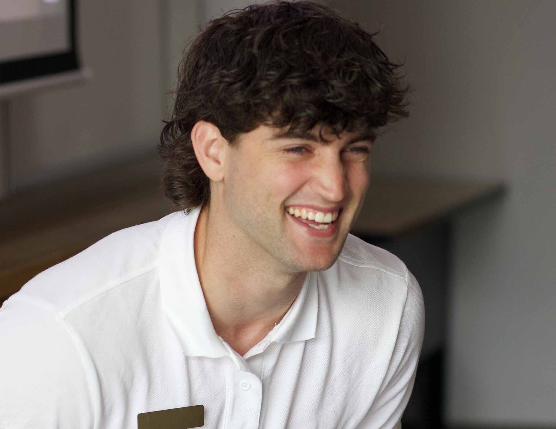 Un jeune homme souriant, vêtu d’un polo blanc, est assis à l’intérieur.