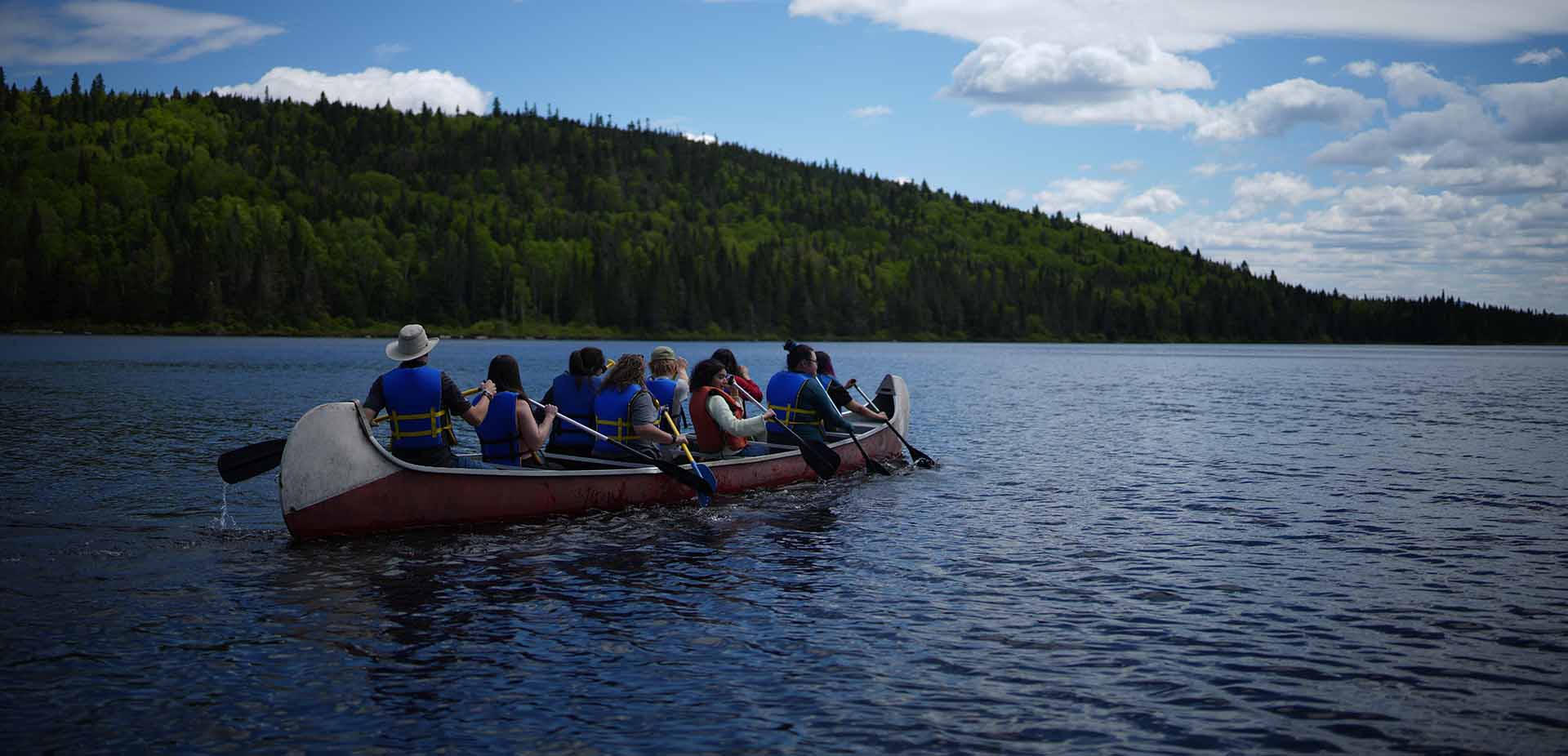 Nine people wearing life jackets paddling a canoe across a calm lake, surrounded by forested hills under a bright blue sky with scattered clouds.