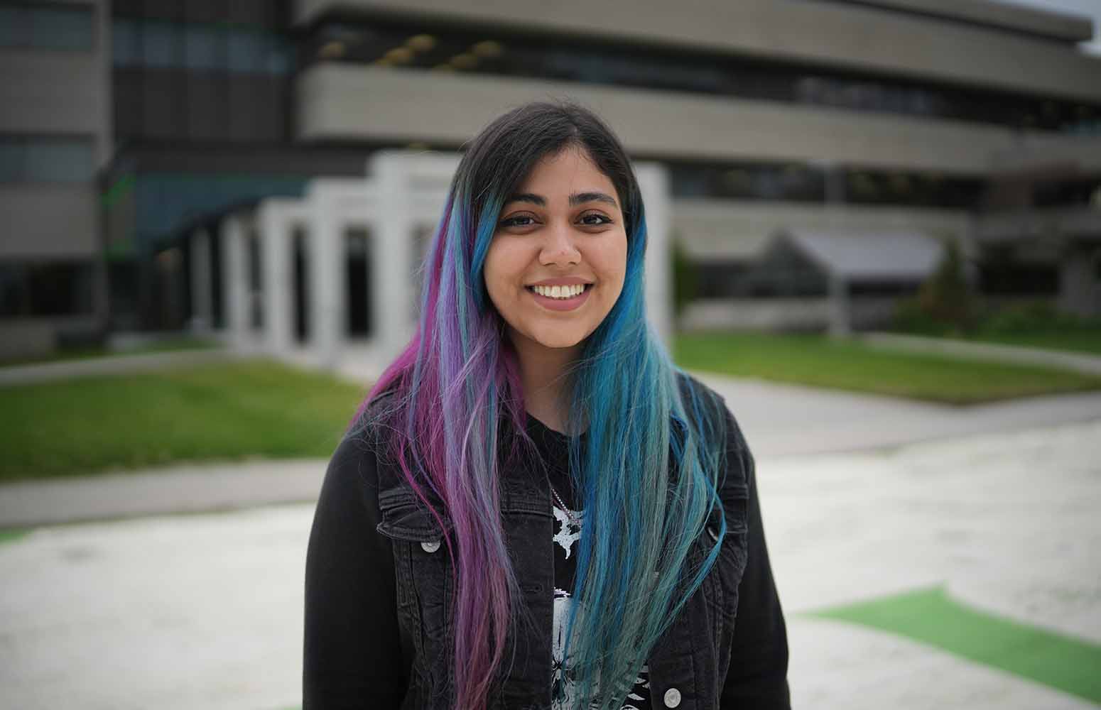 Une jeune femme souriante aux cheveux colorés se tient à l’extérieur, devant un immeuble.