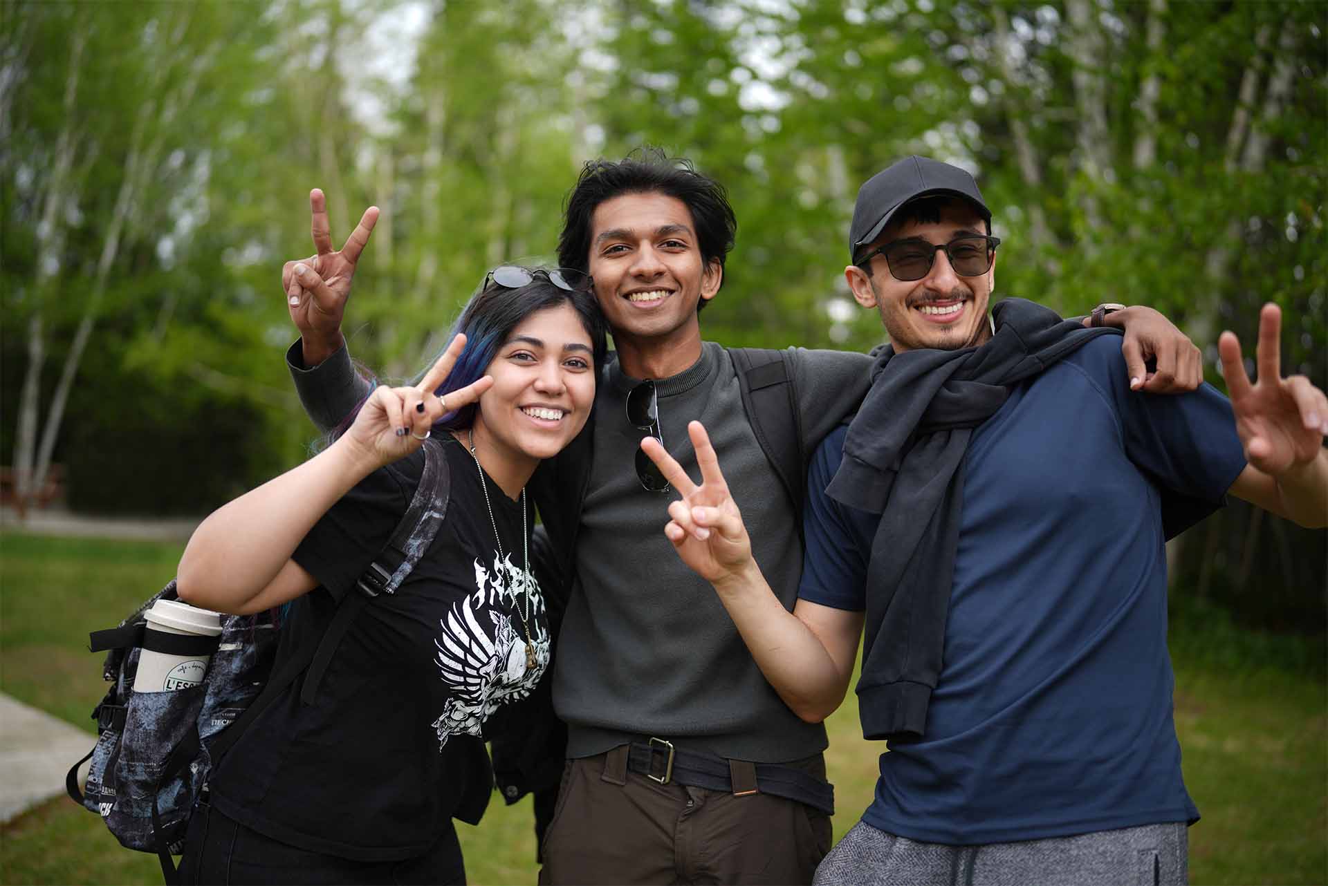 Trois jeunes adultes souriants, munis de sacs à dos, posent ensemble sur un sentier en forêt, en faisant le signe de la paix lors d’une sortie en nature.