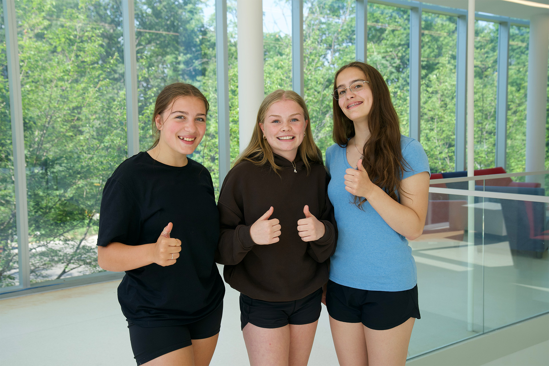 Trois élèves se tiennent côte à côte à l’intérieur. Elles sourient et font un pouce levé devant de grandes baies vitrées donnant sur un espace vert extérieur.