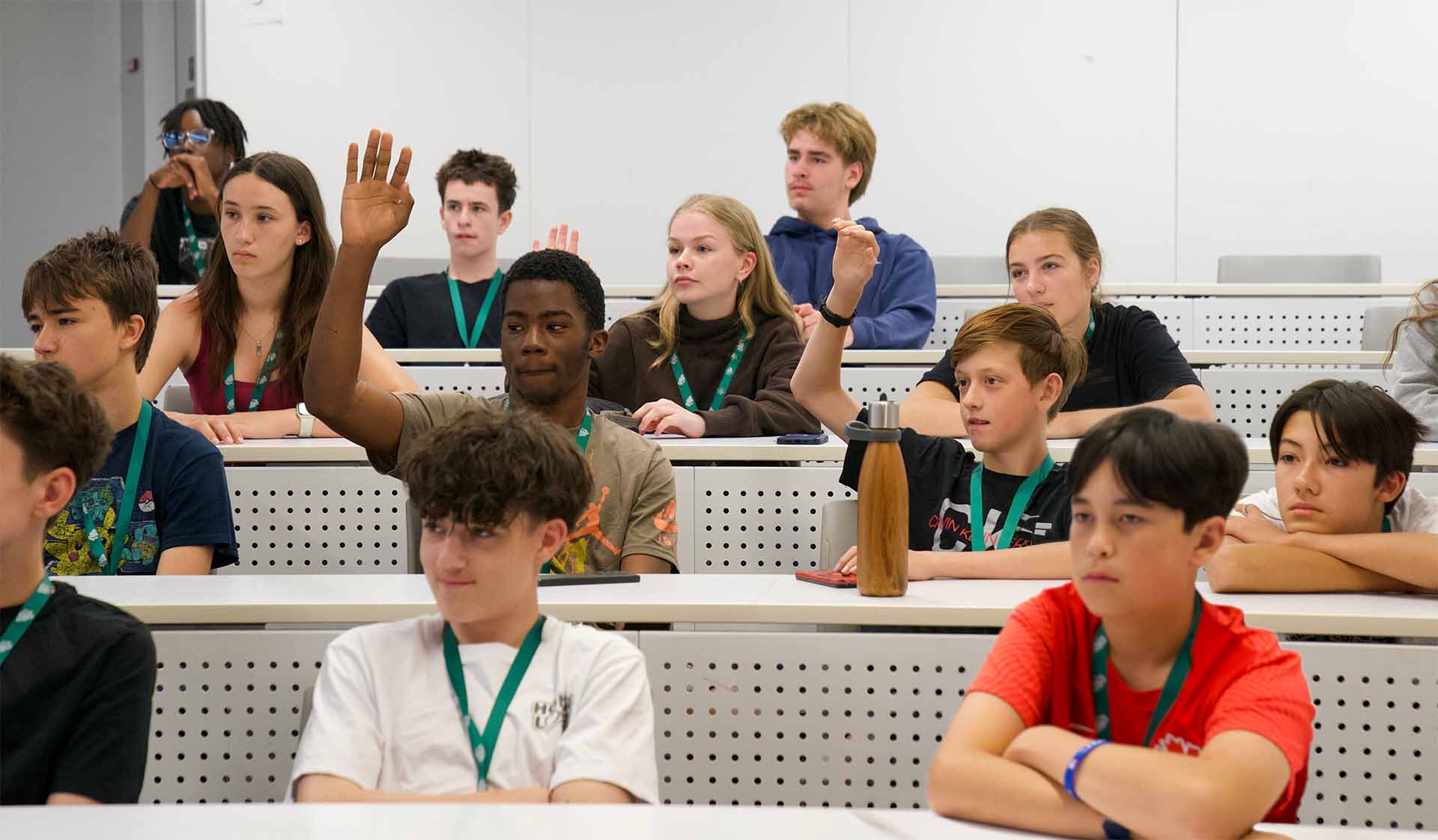 Des élèves sont assis et participent à un cours. Plusieurs lèvent la main pour répondre à une question, tandis que d’autres écoutent attentivement.