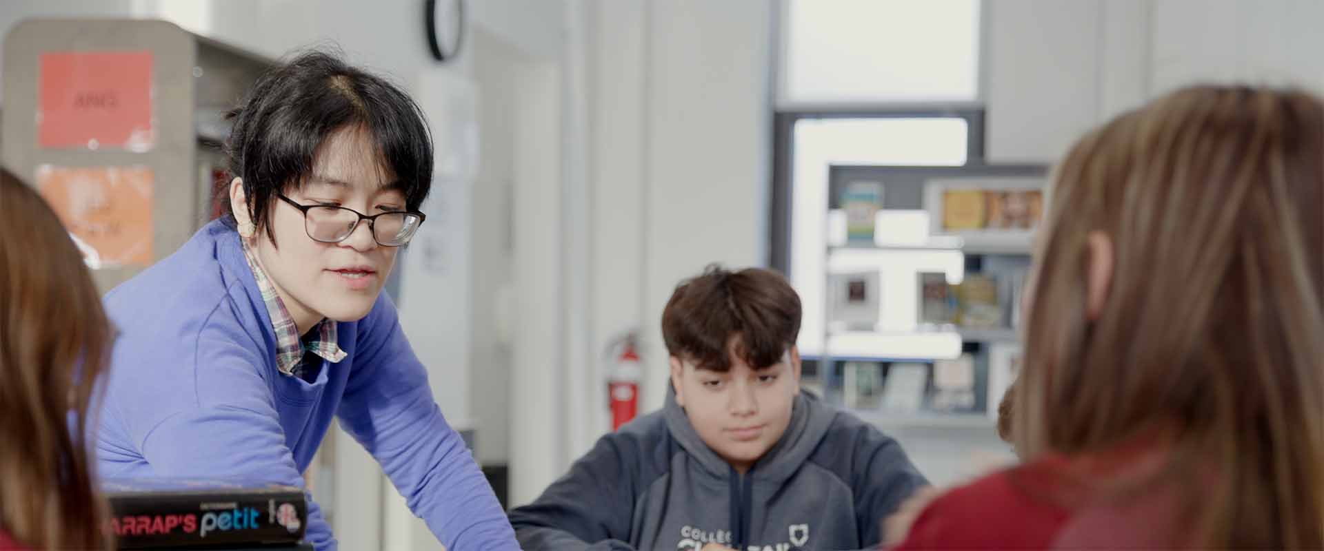 Teacher sitting at a table with four young students, smiling and engaging with them during a hands-on learning activity in a classroom setting.