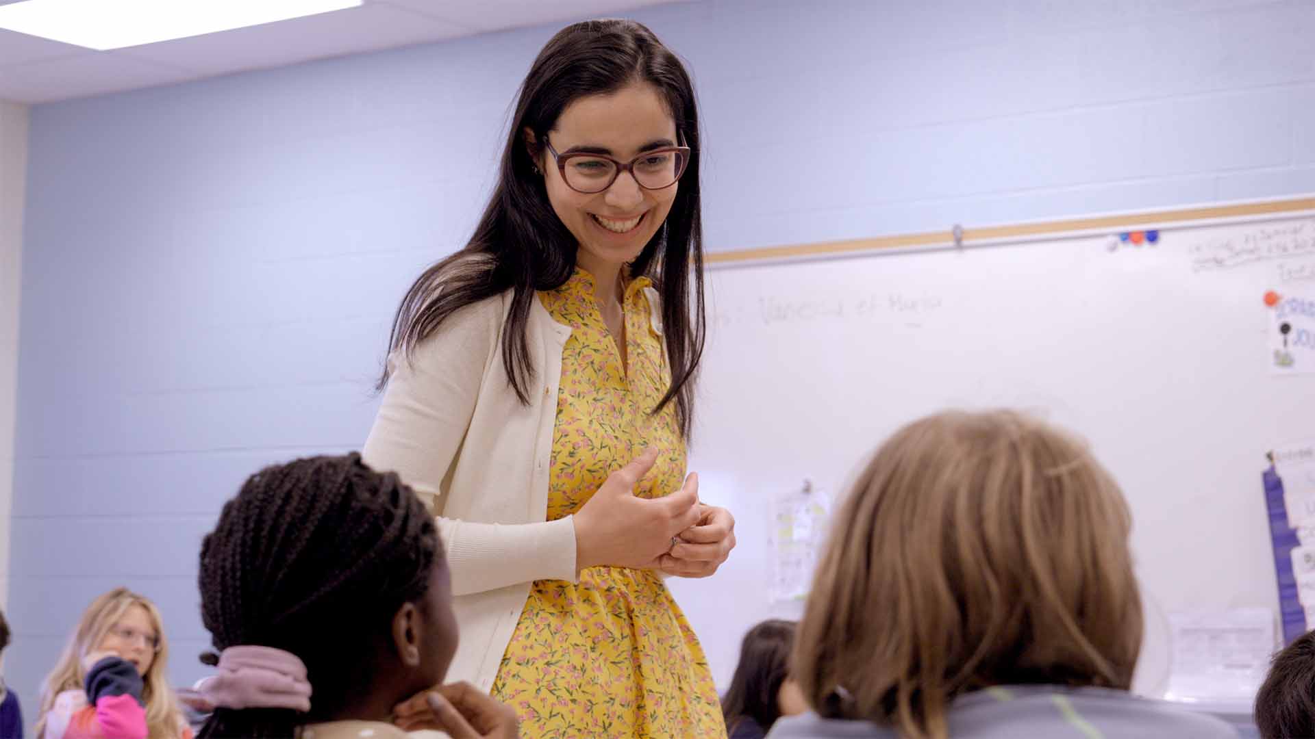 Une monitrice de langues souriante se tient debout dans une salle de classe au milieu d’élèves assis. Elle anime une discussion de groupe tandis que les élèves écoutent et participent.