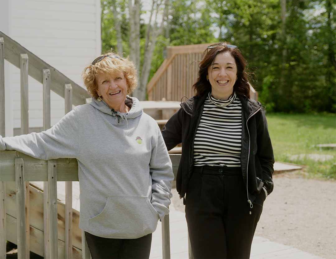 5 – AboutPage_Image18 – FINAL Two women stand outdoors beside an accessible wooden ramp leading to a white building. The women are smiling at the camera with trees and a grassy area in the background.