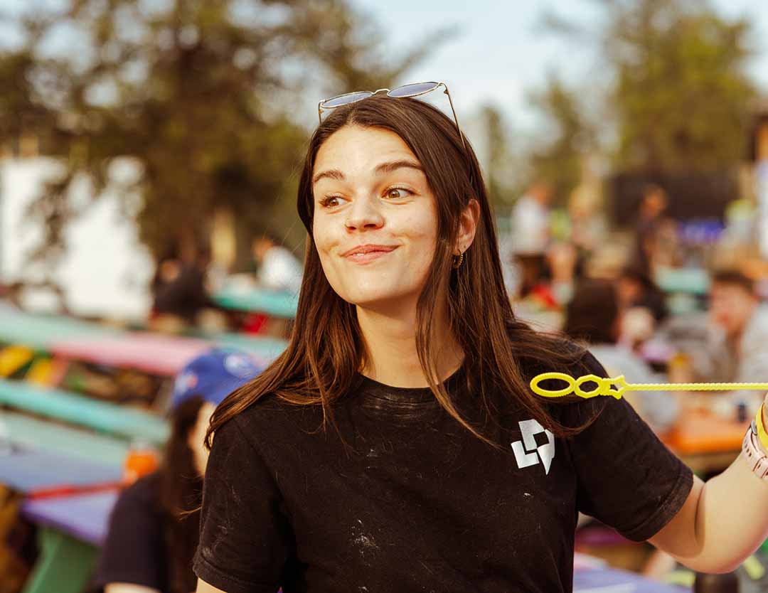5 – AboutPage_Image19 – FINAL (EDITED) A young woman holds a bubble wand at an outdoor gathering, with colourful picnic tables and a blurred crowd in the background.
