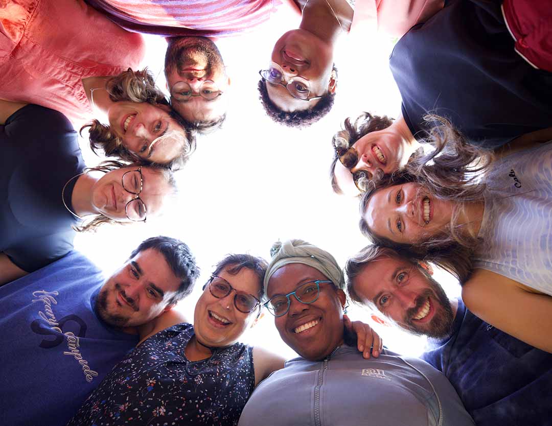 5 – AboutPage_Image20 – FINAL (EDITED) A group of ten people stand in a circle outdoors, looking down toward the camera and smiling, with the bright sky visible in the centre of the group.
