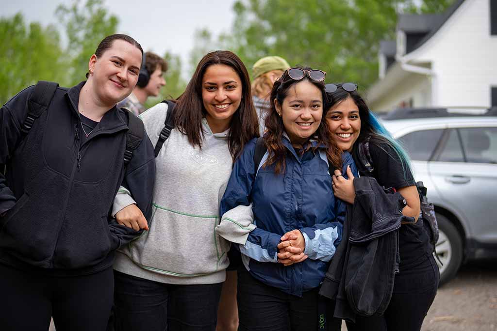 Four youth wearing jackets and
backpacks stand close together
outdoors, smiling at the camera
with trees and parked cars in the
background.