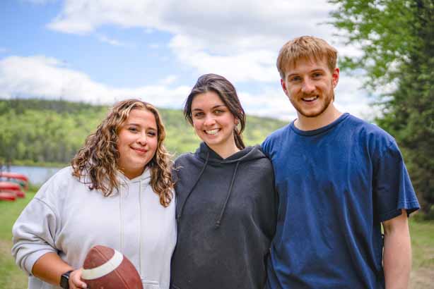Trois jeunes souriants, debout côte à côte à l’extérieur, avec des arbres, des collines et des canots au bord d’un lac en arrière-plan; la personne à gauche tient un ballon de football.