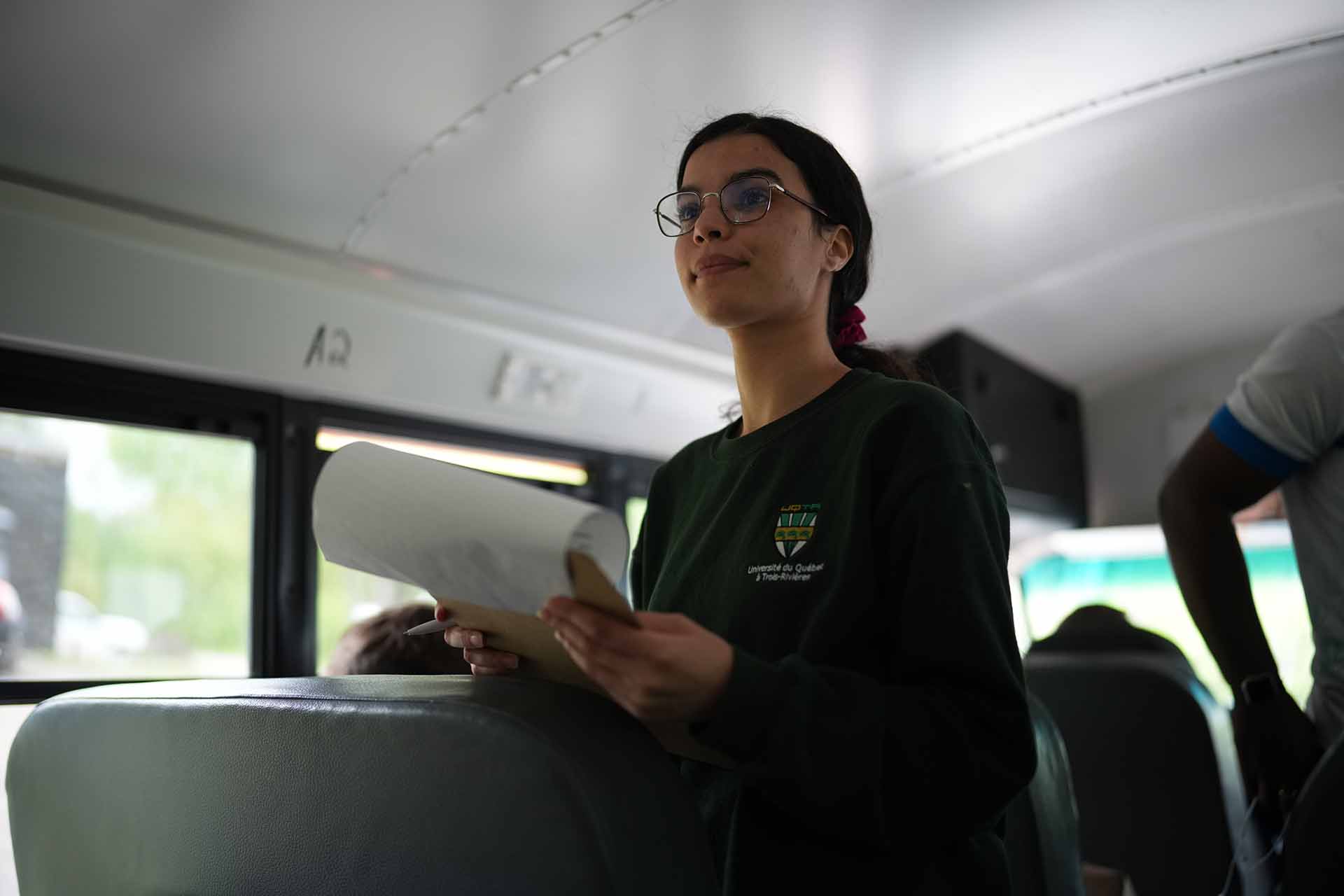 Une jeune femme se tient debout à l’intérieur d’un autobus scolaire. Elle tient des documents et s’adresse aux autres passagères et passagers lors d’une sortie éducative.