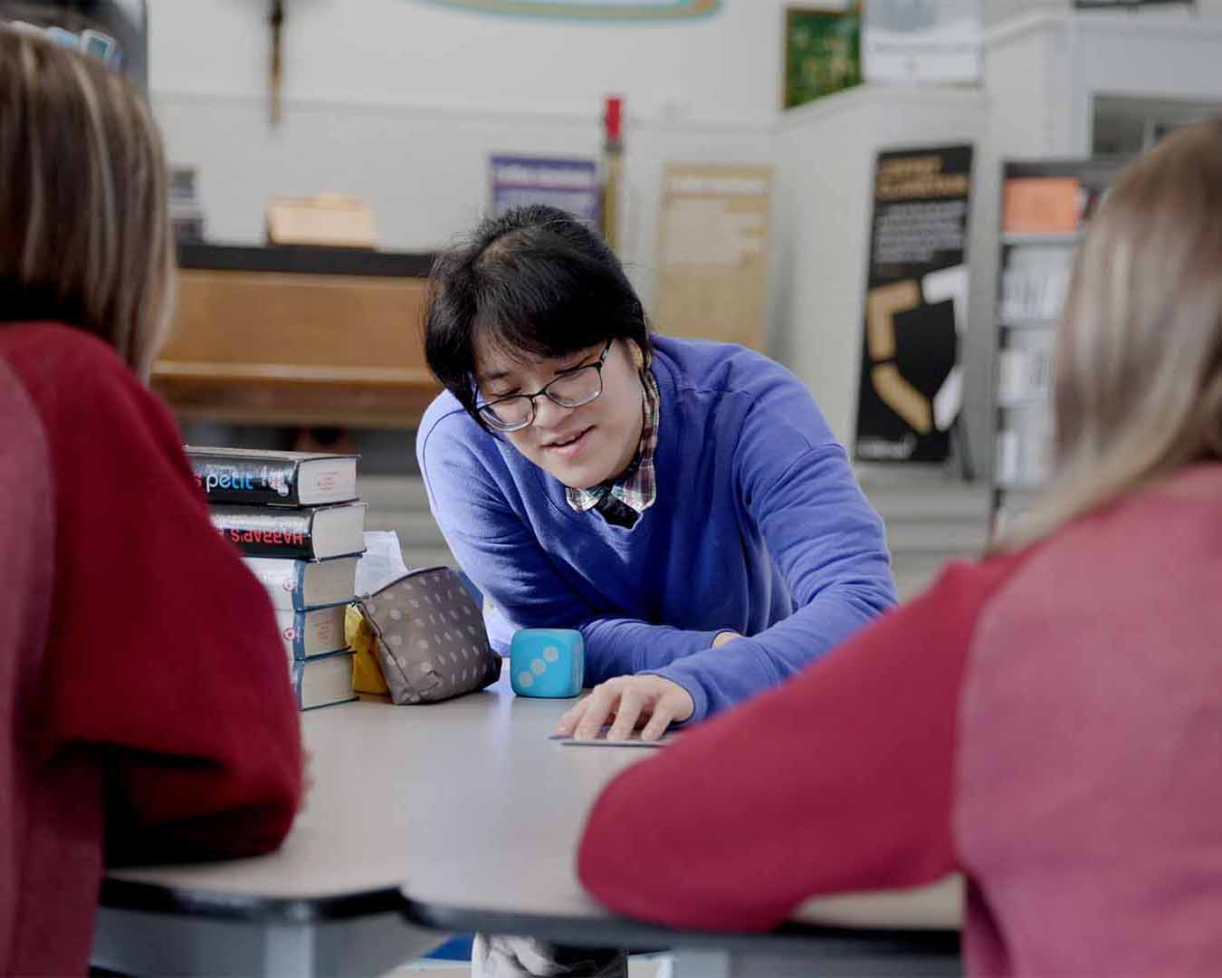 Une monitrice de langues se penche au-dessus d’une table pour présenter une activité à des élèves dans une salle de classe lumineuse. Des livres et du matériel pédagogique sont disposés sur la table. Les élèves sont photographiés de dos et font face à la monitrice.