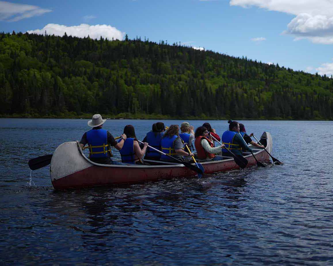 Neuf personnes vues de dos, en gilet de sauvetage, rament à bord d’un canoë sur un lac paisible. Le groupe est entouré de collines boisées, sous un ciel bleu éclatant parsemé de nuages.